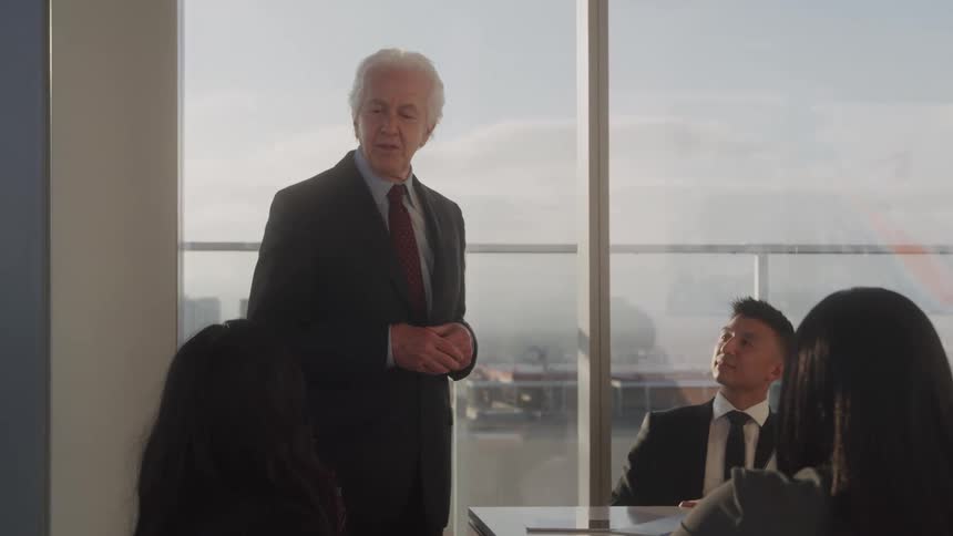 A mature man in a dark suit stands and addresses a group of diverse professionals seated around a conference table in a sunlit room large windows behind them reveal a city skyline with distant buildings and a colorful likely rainbow reflection
