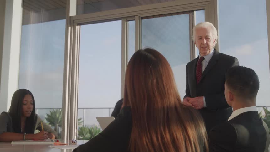 An elderly businessman speaks to younger coworkers while standing in a conference room in an office building during daytime