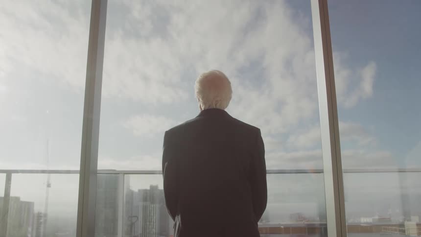 An elderly man looks out a large window while standing in a office building during daytime