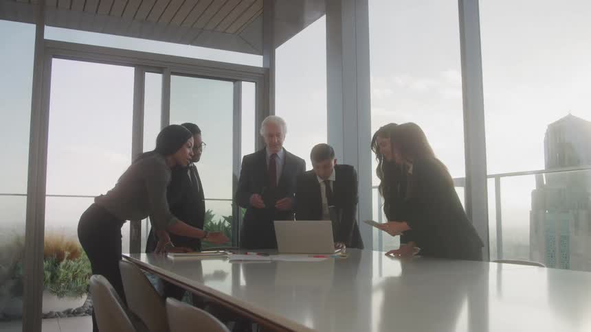 A group of six welldressed business professionals are gathered around a table in a modern office with large windows overlooking a city they appear to be in a discussion with one person pointing at a laptop on the table