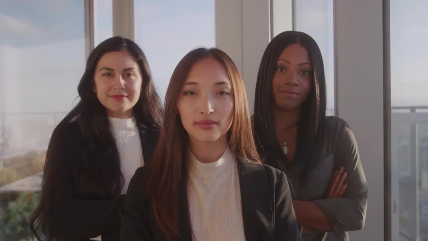 Three diverse professional women stand confidently sidebyside looking directly at the camera with a cityscape visible through windows in the blurred background