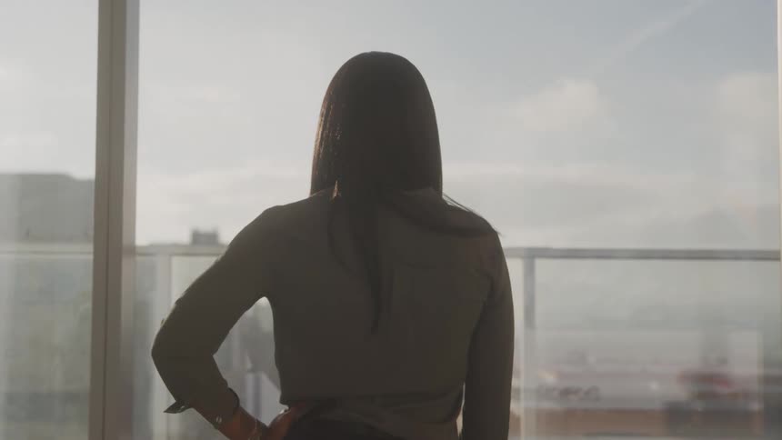The video shows a person with long dark hair seen from behind standing in front of a large window they are looking out at a cityscape with buildings and what appears to be an airport or port visible in the distance under a bright sky