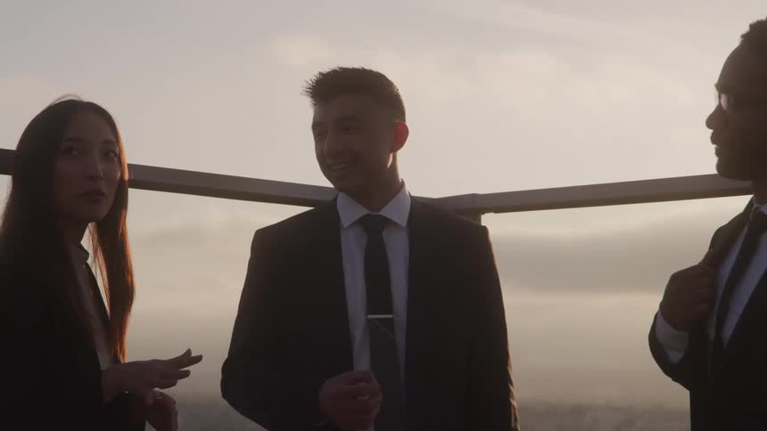 The video shows three individuals two men and one woman in professional attire engaged in a conversation on what appears to be a rooftop or highrise balcony the setting sun casts a warm golden glow on them suggesting its either sunrise or sunset