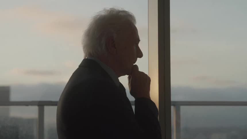 An older gentleman gazes thoughtfully out a highrise window at dusk his hand resting on his chin as he reflects in quiet contemplation