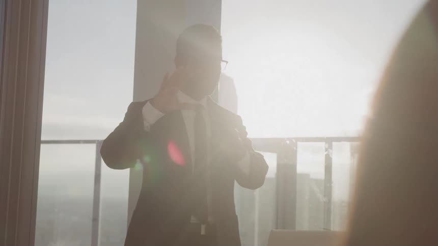 A man in a suit and tie stands in front of a large window with bright sunlight streaming in from behind him he is seen adjusting his suit and glasses while speaking to someone offcamera