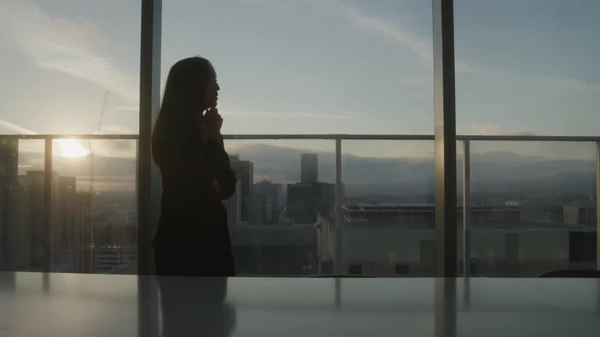 A woman stands silhouetted in a highrise office at sunset speaking on the phone as golden light spills across the city skyline behind her