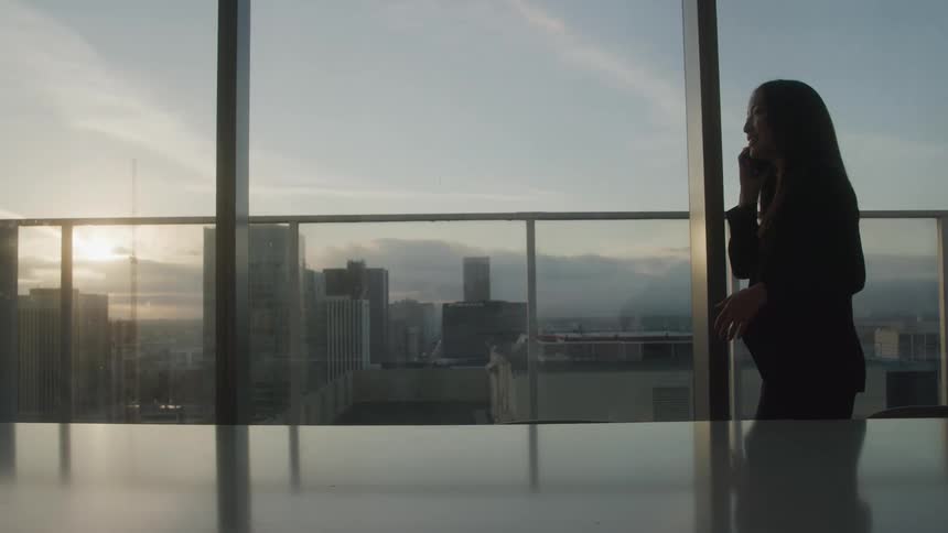 A woman is silhouetted against a large window with a cityscape view at sunset she is pacing and talking on her phone
