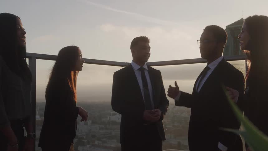 A group of professionals in formal attire engage in lively conversation on a rooftop at sunset with warm city light casting a golden glow on their exchange
