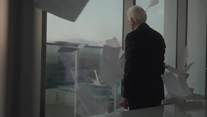 An elderly man with gray hair in his office at sunset tosses all the papers into the air causing them to fly abruptly as he stares out the window