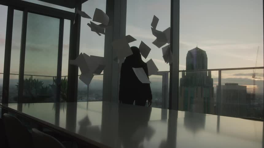 The video shows a silhouetted figure standing in front of large windows looking out at a city skyline with a long table in the foreground several sheets of paper are seen falling around the figure landing on the table a filmsupply watermark