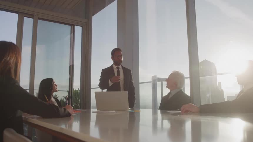 A group of people sits at a conference table and watches a man speak