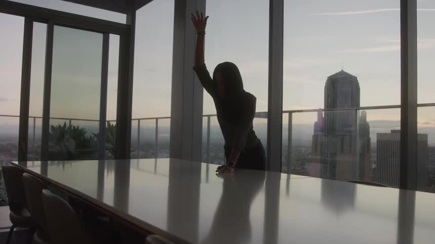 A woman throws paper documents in the air after reading them at a table in an office building at sunset