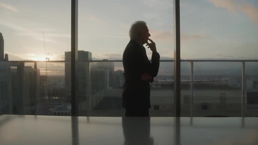 An older man in a dark suit stands silhouetted against the soft glow of a city skyline at sunset gazing out from a highrise window while thoughtfully holding his hand to his mout