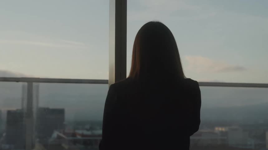 A woman stands in front of a large window that reveals a city skyline holding a phone to her ear as she speaks