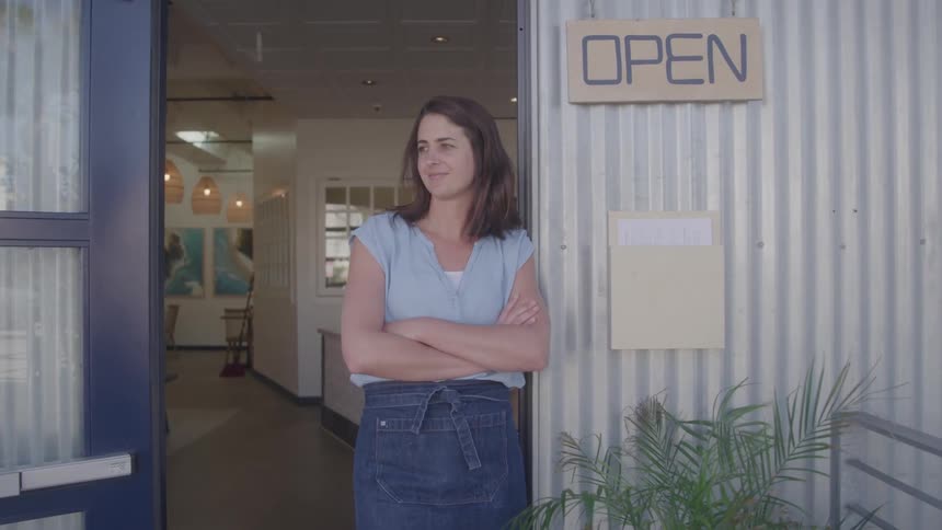 A woman in a denim apron stands proudly in the doorway of her small business arms crossed beneath an open sign