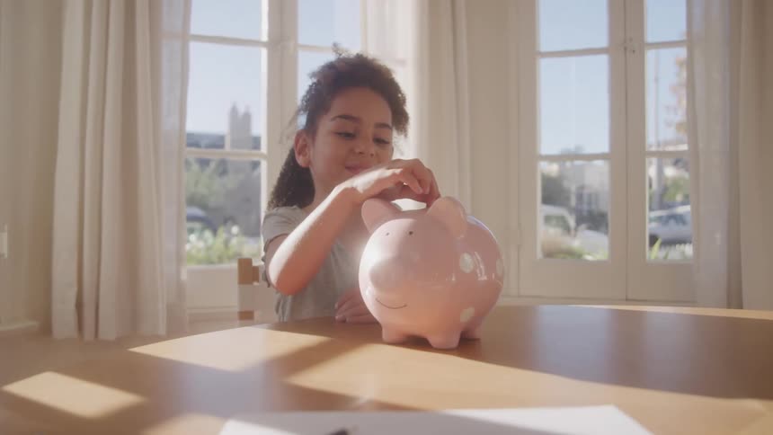 A girl child drops a coin into a pink piggy bank on a table indoors during daytime