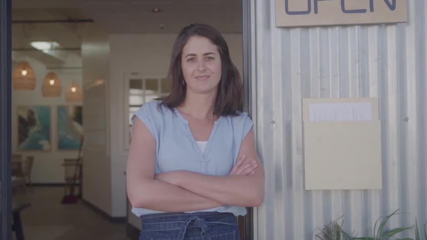 A smiling woman with dark hair and a denim apron stands confidently with her arms crossed in the doorway of a restaurant featuring wicker light fixtures and artwork inside