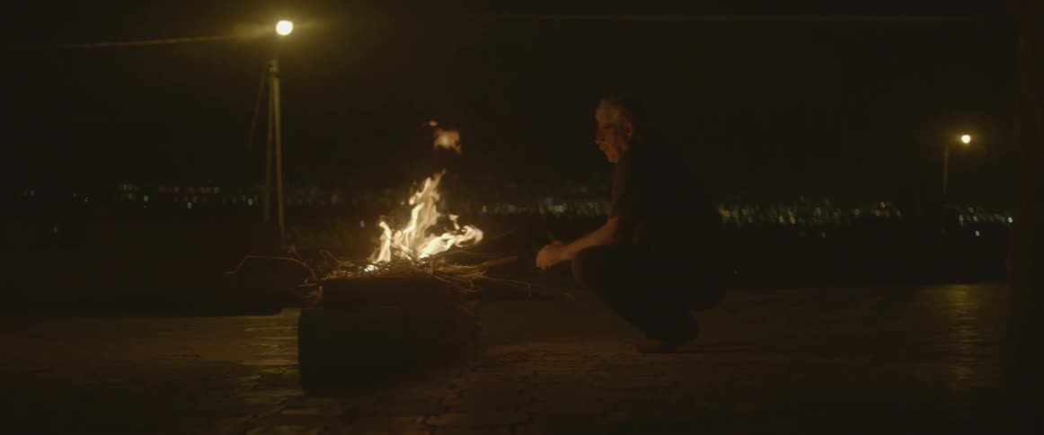 A man squats near a burning fire at night