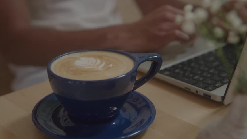 A rich creamy latte in a deep blue cup sits invitingly beside a person typing on a laptop blending focus with comfort in a cozy workspace