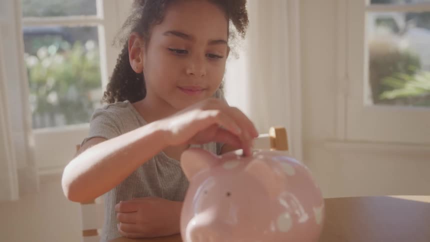 A young girl with curly hair is shown smiling gently as she inserts a coin into a pink piggy bank on a wooden table sunlight streams in from a window behind her illuminating the scene