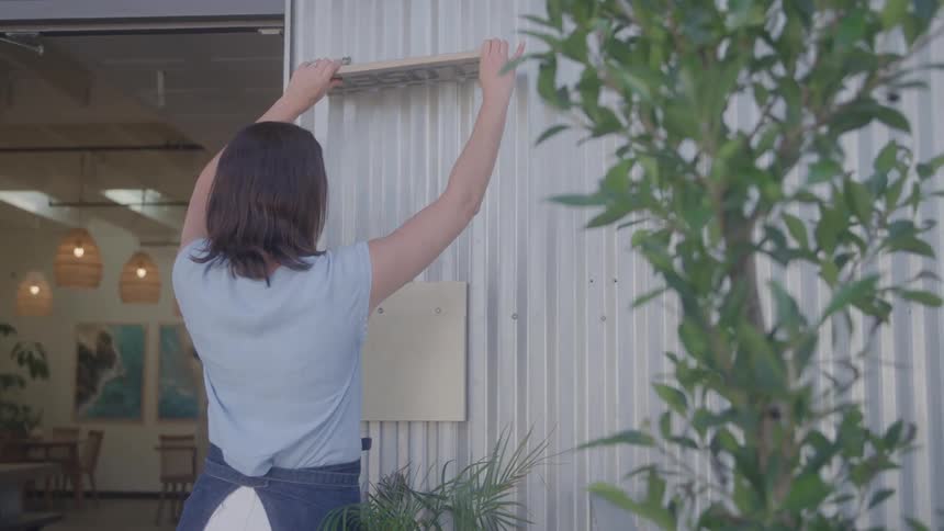 A woman with her back to the camera is seen hanging an open sign on the exterior wall of a building lush green foliage is visible on the right side of the frame suggesting a pleasant outdoor setting for the establishment