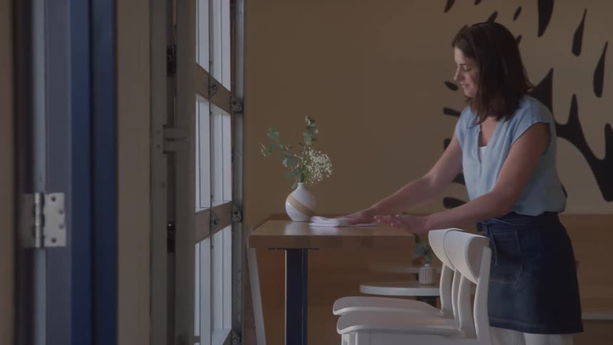 A woman with dark hair wearing a light blue top and a dark apron is seen wiping down a wooden table in what appears to be a cafe or restaurant a vase with flowers is on the table and theres a large window or door on the left side of the frame