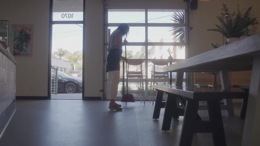 The video shows a person sweeping the floor of what appears to be a cafe or shop with tables and chairs visible in the foreground through the large windows in the background a car can be seen parked outside