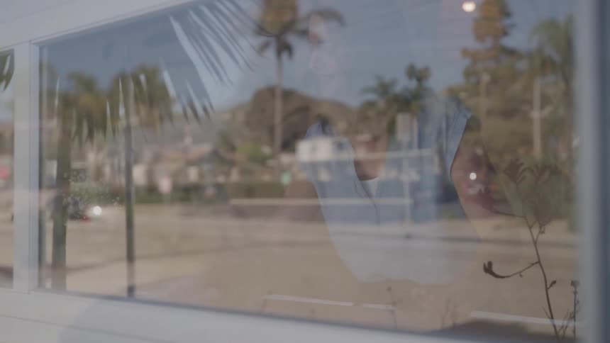 A young woman is seen through a glass window cleaning a table inside a restaurant with a rag at midday