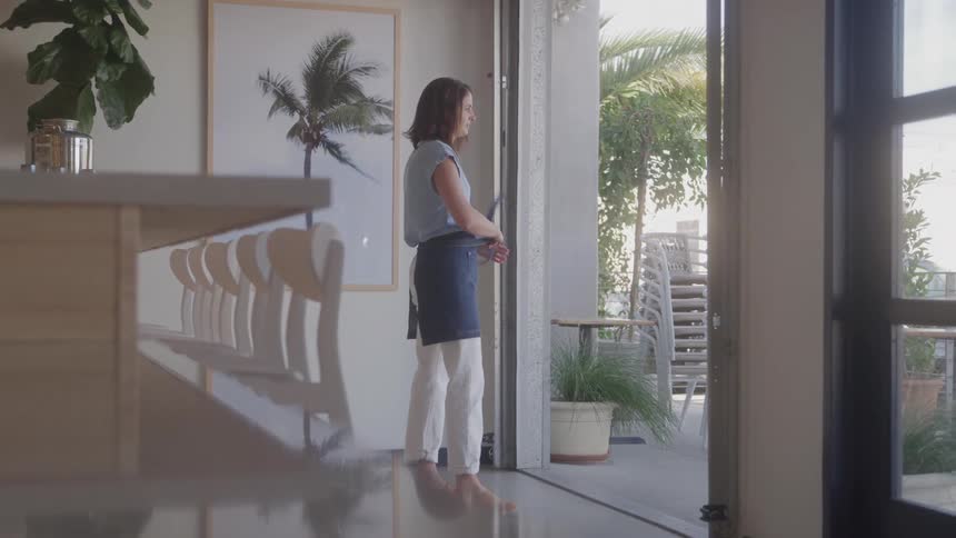 A woman stands near a large open doorway looking out towards an outdoor patio area with palm trees she is wearing a lightcolored top and what appears to be a dark apron or skirt suggesting she might be working in a cafe or restaurant
