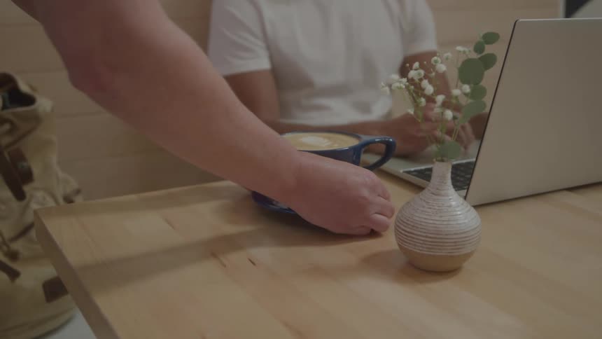A persons hands are seen placing a blue cup of latte art coffee onto a wooden table in the background another person is sitting at the table working on a laptop with a small vase of flowers nearby