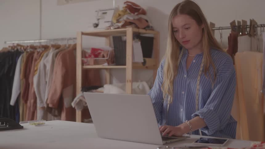 A young female fashion designer uses a laptop at a table in a workshop