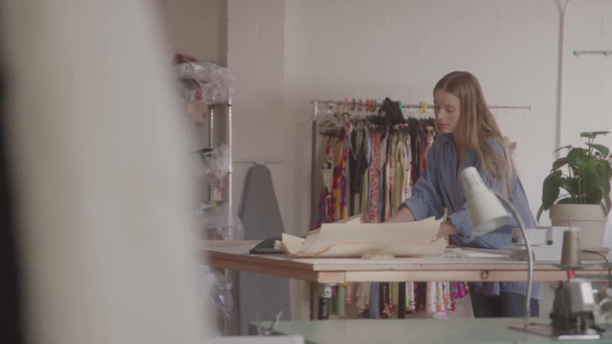 A young woman with long blonde hair wearing a striped blue shirt is standing at a large wooden table in a welllit room she is carefully unfolding what appears to be a paper pattern or large document