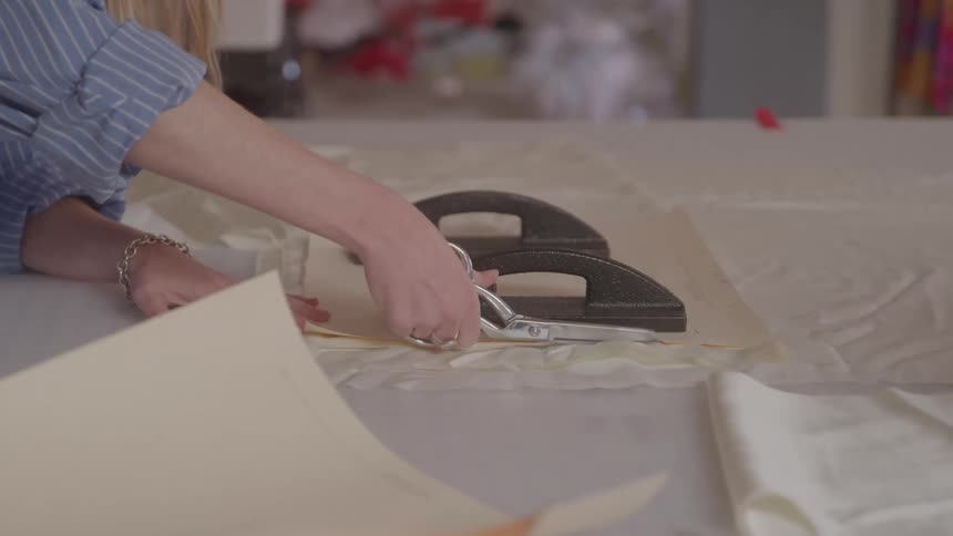 A person uses fabric scissors to cut along a pattern pinned under a heavy black weight on a large table in a sewing or design workspace