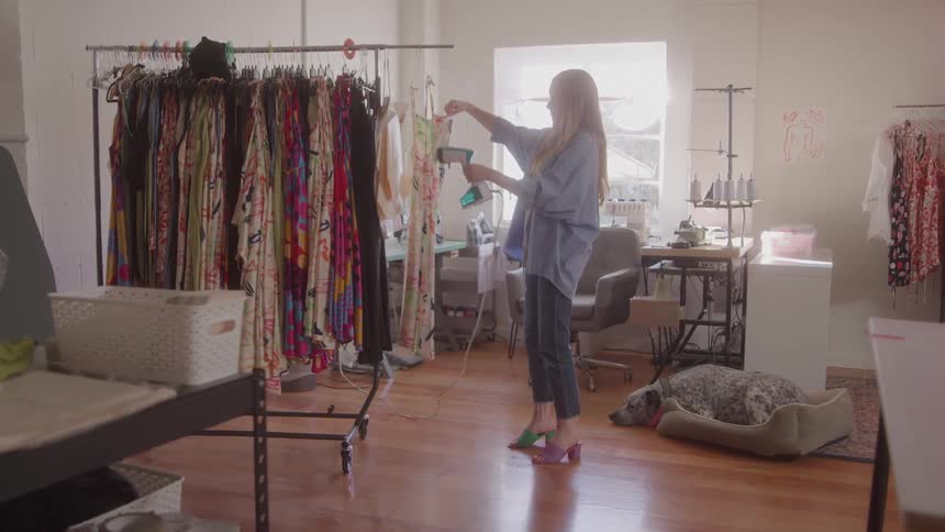The video shows a woman standing in a room filled with clothes racks using a handheld steamer on a white dress a dog is peacefully sleeping in a bed on the floor adding a domestic touch to the scene that appears to be a fashion studio or a retail space