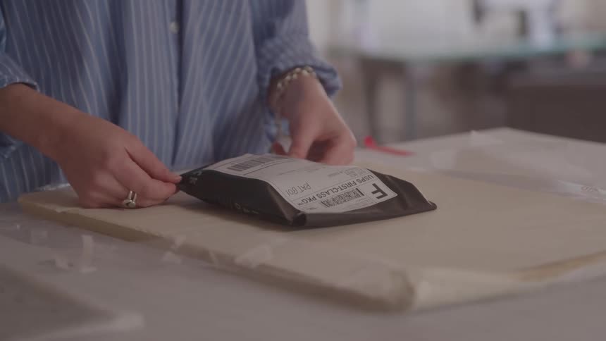 A person wearing a blue striped shirt and a ring is shown carefully handling a small darkcolored package on a lightcolored surface the package appears to be an envelope or soft mailer