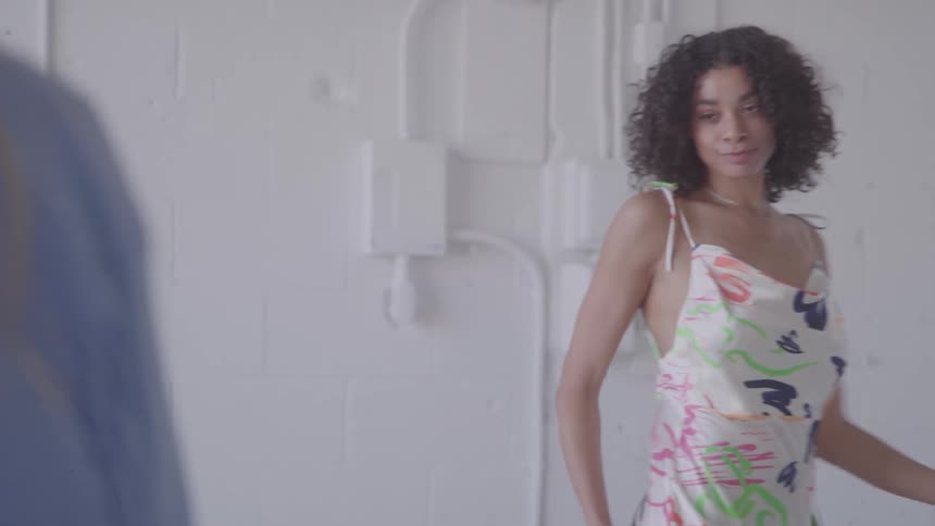 A woman with curly hair wearing a colorful patterned dress is posing for a photo another person partially visible in the foreground is holding a smartphone and taking the picture