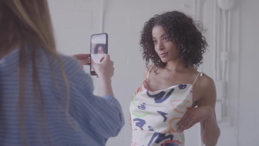 A woman with dark curly hair wearing a patterned slip dress poses in front of a plain white wall another person with lightcolored hair is seen from behind holding up a smartphone to take her picture