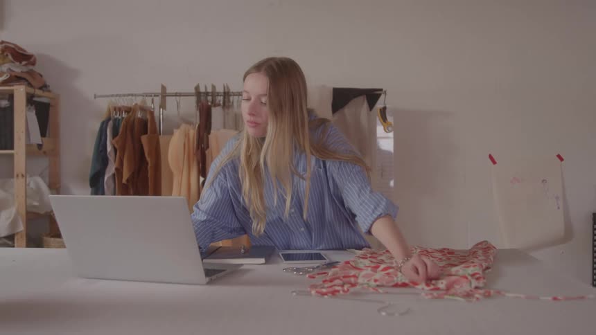 A woman designs fabric in a studio with a laptop and clothes