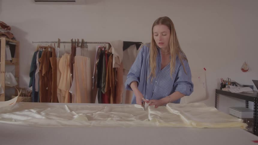 A woman with blonde hair wearing a striped blue shirt is meticulously cutting fabric on a large table in the background a clothing rack filled with garments is visible