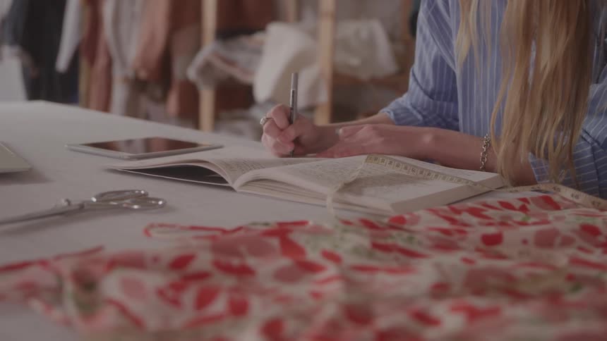 The video shows a person with long blonde hair wearing a striped shirt writing in a notebook at a desk on the desk there is also a tablet scissors and a piece of fabric with a floral pattern a filmsupply watermark is visible across the center of