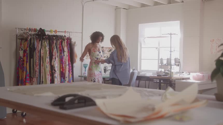 A woman takes measurements of another woman in a tailoring shop at midday with a hanger full of dresses beside them and a sewing machine on the right side