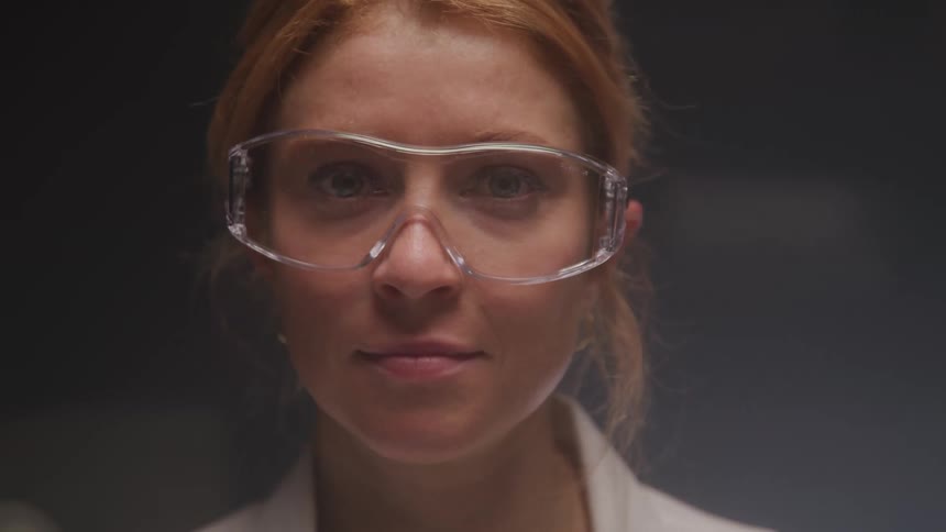 A closeup shot features a woman with reddishblonde hair pulled back wearing clear safety goggles and a white lab coat she looks directly at the camera with a calm expression