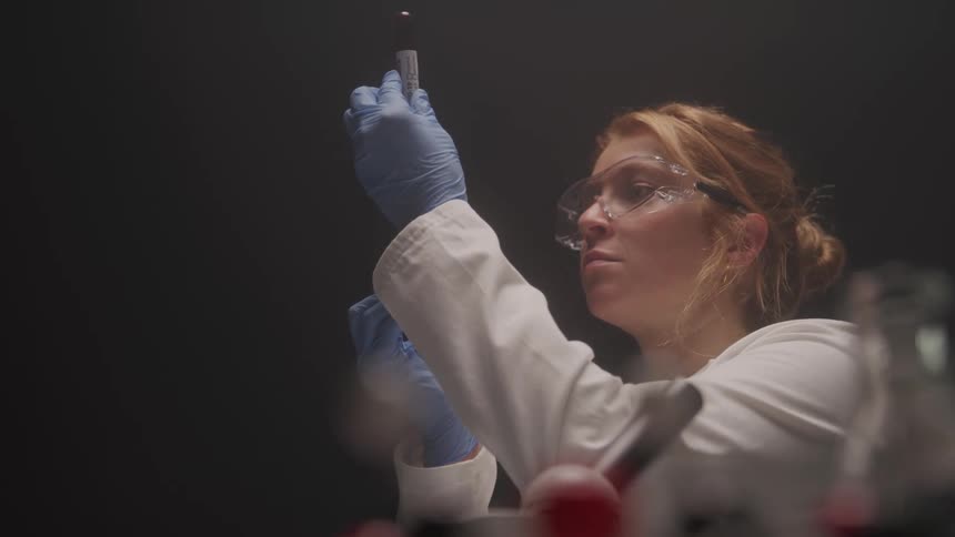 A woman wearing safety glasses and blue gloves holds a small test tube with a clear liquid and a drop at the top looking intently at it in a dimly lit laboratory