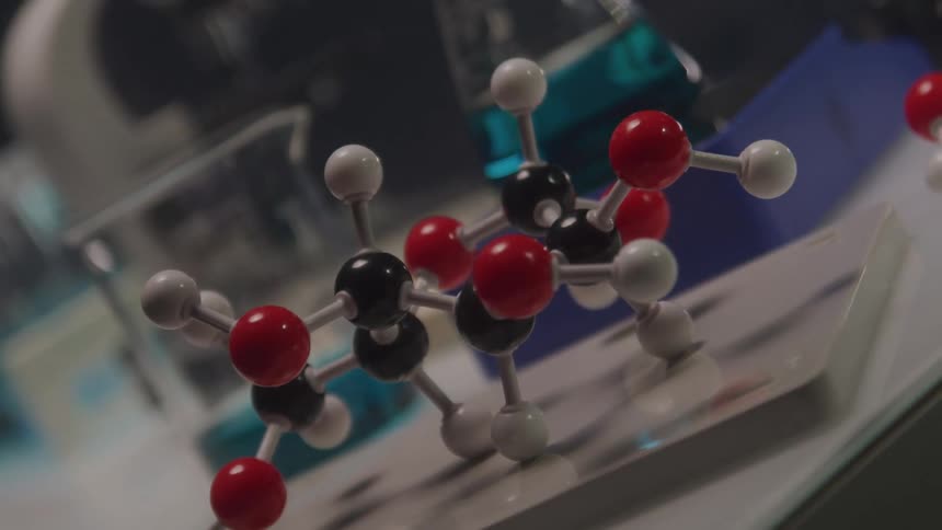 A detailed molecular model with red black and white spheres connected by rods sits on a white surface with blurred beakers of blue liquid and other lab equipment in the background