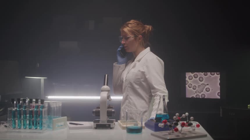 A focused scientist in a white lab coat speaks on the phone while surrounded by test tubes a microscope and molecular models in a dimly lit laboratory