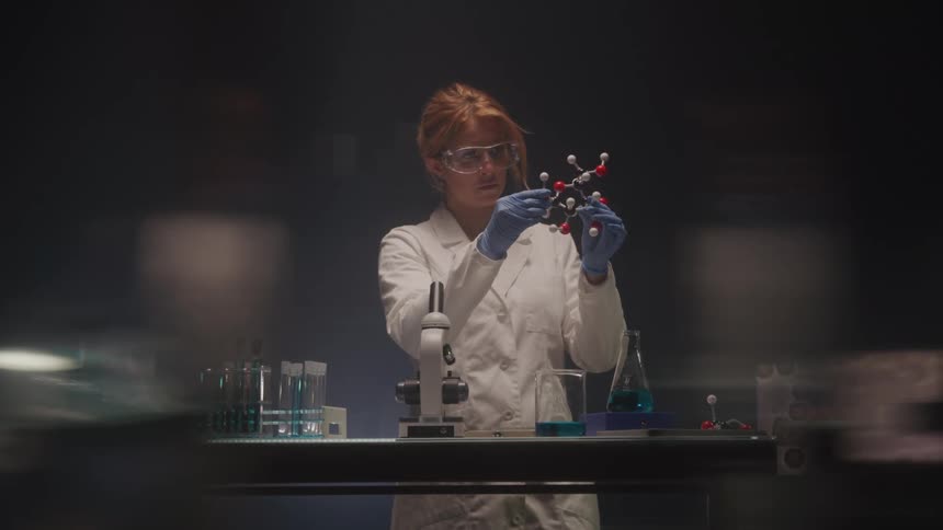 A lab technician wearing goggles and a lab coat looks at a molecule through a microscope with beakers on the table in front