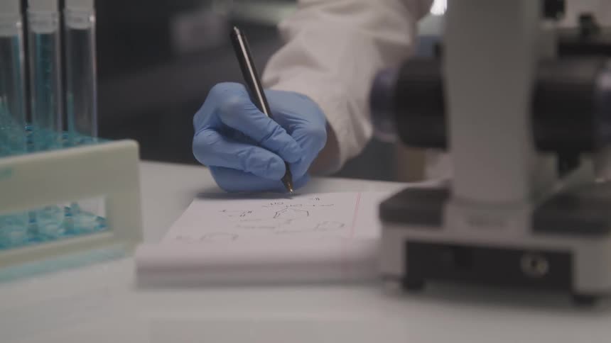 A gloved hand writes notes beside a microscope in a laboratory setting