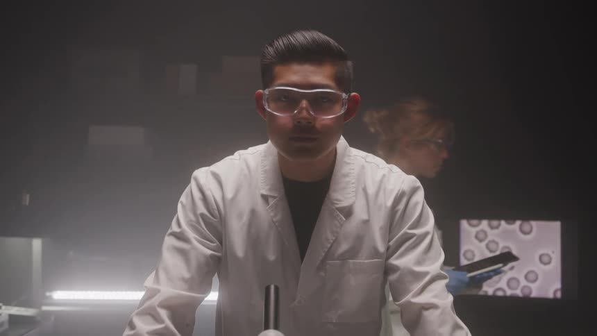 The video features a man in a lab coat and safety goggles looking directly at the camera with a serious expression in the background another person in a lab coat can be seen moving and a screen displays what appears to be microscopic images of cells