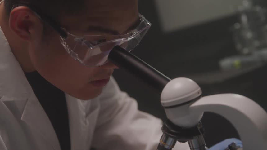 A person in a white lab coat and safety goggles is intensely looking through a microscope the setting appears to be a laboratory