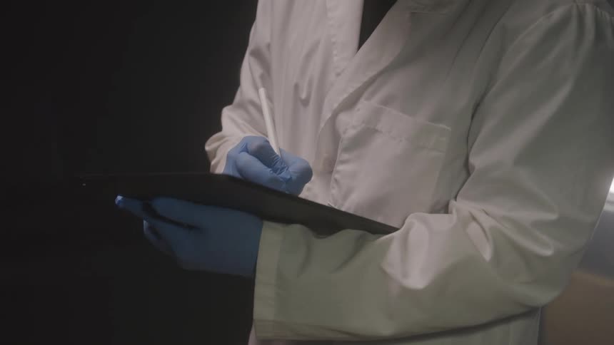 Person in white coat and gloves holding a clipboard and taking notes